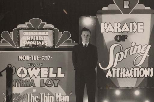 Black-and-white photograph of man standing in front of a theatre placard. The movies on display include The Thin Man (1934), starring William Powell and Myrna Loy; and The Plainsman (1936), starring Gary Cooper and Jean Arthur. The display also announces that the "Imperial Hawaiians," a group of male musicians, were scheduled to perform onstage. The "R" at the top of the display as well as the architectural details at the top of the photograph point to the location of this photo as the Rylander Theatre, in Americus Georgia. 