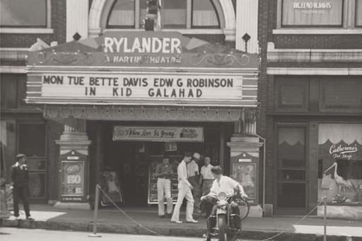 A black-and-white photo of the facade of the Rylander Theatre in Americus, Georgia. The marquee reads "Mon Tue Bette Davis Edw[ard] G. Robinson in Kid Galahad." A young man sits on top of a motorcycle as he looks back at the theater, and pedestrians walk along the sidewalk in front of the theater.