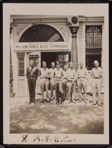 A photograph of a group of servicemen standing in front of the building headquarters for the United States Eighth Air Force Base Command in Savannah, Georgia. A sign on the building reads "Headquarters Eighth Air Force Base Command." The back of the photograph reads: "Early 1942 Savannah Ga."