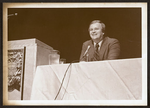 Image courtesy of University of Georgia Alexander Campbell King Law Library Archives & Special Collections. Photograph (front side) of Max Cleland, then the U.S. Veterans Administration director, seated at a dais onstage at the University of Georgia School of Law, where he delivered the commencement address on June 6, 1978. Transcribed from the back of the photo: "Max Cleland, graduation speaker."